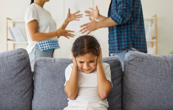 Child covering ears during father and mothers conflict to ignore angry words at home. Frustrated girl with trauma and stress from parental arguments and quarrel sitting on sofa in living room