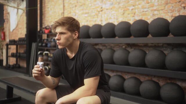 Focused young athlete in black athletic wear consuming protein bar after intense workout in industrial gym