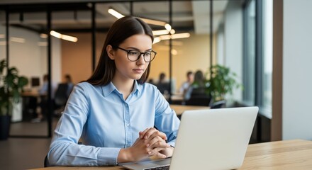 A serious, professional young woman wearing glasses and a blue shirt works on a laptop with clasped hands in a bright, modern open-plan office setting.