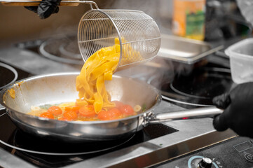 Chef pours cooked pasta into pan with cherry tomatoes and garlic on stovetop. Steam rises. Modern kitchen, cooking in progress, vibrant Italian dish preparation.
