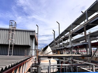 Fototapeta premium Wide-angle view of a vast industrial complex featuring a labyrinth of overhead pipes, metal scaffolding, and corrugated steel buildings under a bright, cloudy sky, symbolizing heavy industry.