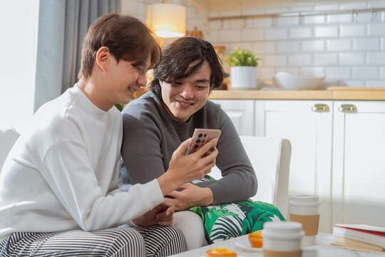 Asian LGBTQ couple shares intimate moments together on sofa, browsing cherished travel photos and precious memories on smartphone while enjoying quality weekend bonding time at comfortable home