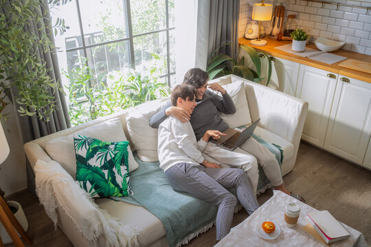 Asian LGBTQ male couple enjoys quality bonding time together using laptop and smartphone for social connections while relaxing on comfortable living room sofa, strengthening relationship through share
