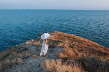 A girl in a big white hat walks on the mountain against the background of the sea and the sunset. Vacation in the fresh air. Joy, travel. High quality photo