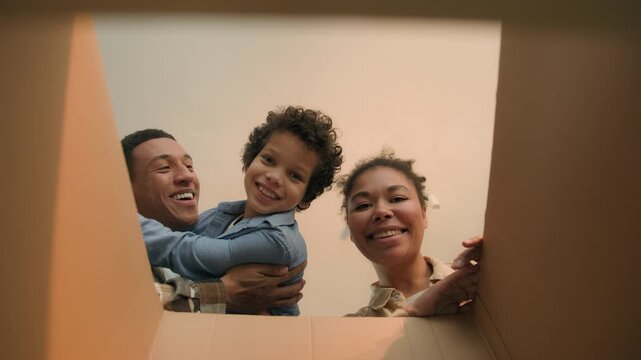 Smiling African American family ethnic parents cuddling son opening cardboard box parcel looking inside closing package lovely bonding mother father with child enjoying delivery service moving day