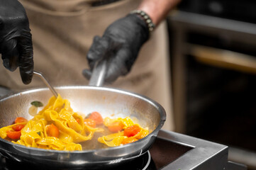 Chef cooking tagliatelle pasta with cherry tomatoes in a hot pan. Steam rising, black gloves, beige apron, professional kitchen atmosphere.
