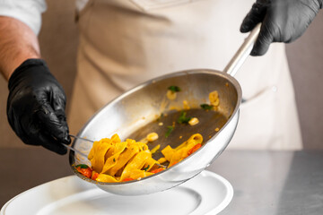Chef plating tagliatelle pasta with cherry tomatoes, garlic, and herbs from frying pan onto white plate. Culinary preparation in kitchen setting, close-up, food styling concept.