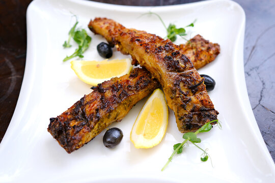 A delicious and beautifully presented dish of two grilled fish, garnished with fresh lettuce, cucumber slices, and sliced red chili peppers, served on a white plate with a red checkered.