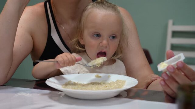 Blonde toddler, nestled with parent, tenderly learns to spoon-feed porridge during heartwarming morning meal, highlighting loving family bond.