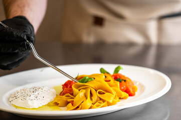 Chef plating tagliatelle pasta with cherry tomatoes, basil, and burrata cheese in a professional kitchen. Gourmet Italian dish styled with precision and care.