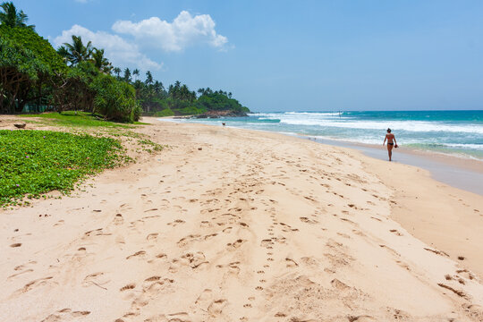 View of golden sand meeting turquoise waves under a vast blue sky, with footprints leading toward a lone figure walking on the shore, Sri Lanka.