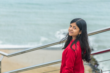 A teen in a red jacket stands on a railing overlooking the ocean