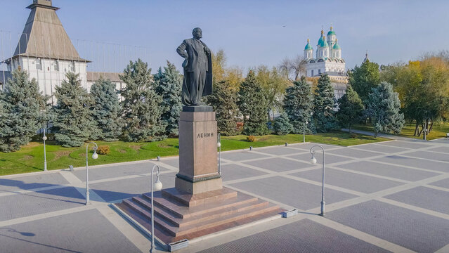 Lenin monument on Lenin Square in Astrakhan, Russia, with historic Kremlin tower, pine trees, and Assumption Cathedral on a sunny autumn day.

