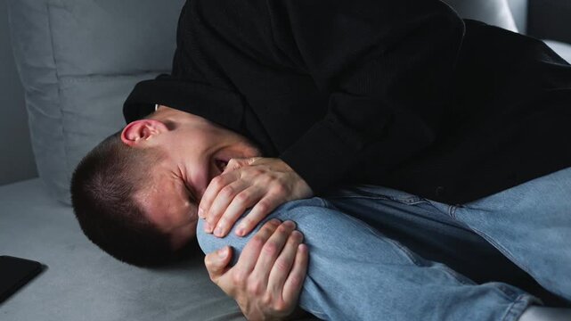 A man holds his injured knee with his hands.
Pain in the joints of his legs.
A man with a bruised kneecap sitting on a gray sofa in a room.