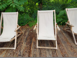 Three empty wooden deck chairs with white canvas seats, placed on a rustic wooden patio surrounded by dense green foliage. The image evokes themes of rest, nature, and summer vacation.