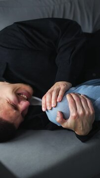 A man holds his injured knee with his hands.
Pain in the joints of his legs.
A man with a bruised kneecap sitting on a gray sofa in a room.