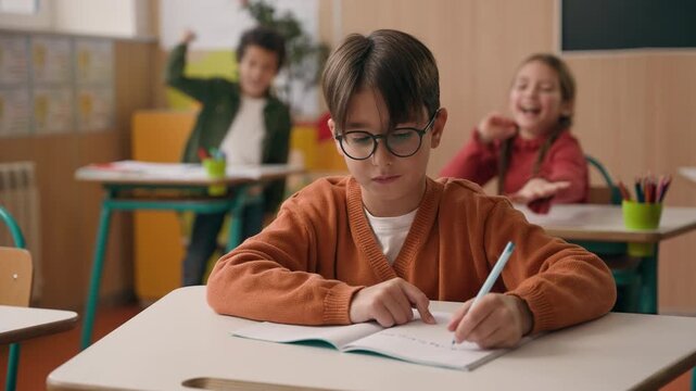 Caucasian boy in school class writing in copybook learning studying ignoring classmates play noisy children schoolgirl girl kid schoolboy child throwing crumpled paper balls laughing mocking bullying