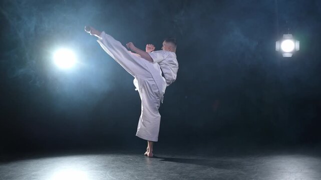 Strong man fighter in white kimono for karate martial arts with black belt preparing for fight training in black background. Man training moves punches kicks practicing shadow fight ready for sparring