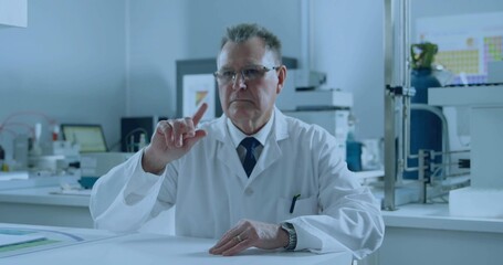 Scientist wearing lab coat pointing at periodic table on monitor in research lab, with gas cylinder