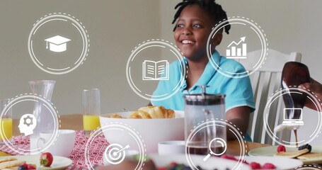 Smiling Black boy sitting at dining table with breakfast and digital learning icons, copy space