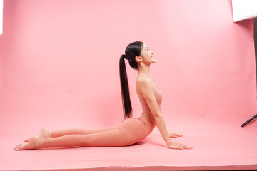 A young woman does the cobra pose on a pink background looking upward smiling brightly