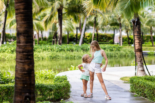 Two adorable caucasian little girls smiling outdoor at hot summer day. Sisters posing in tropical garden with palm trees