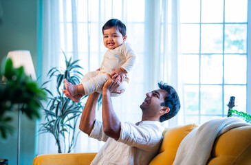 Indian Playful father and baby boy smiling while playing on living room sofa at home