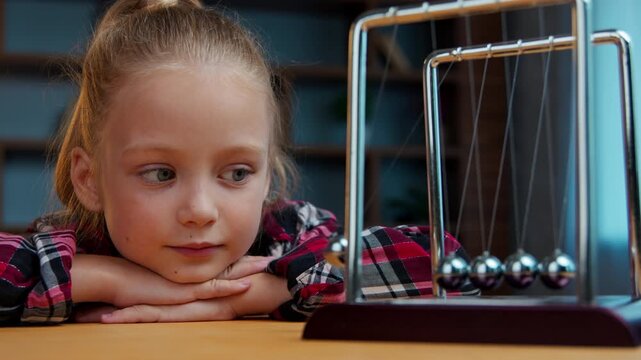 Caucasian little girl smiling child kid daughter cute baby looking at swinging metal steel balls law kinetic energy hypnotized game relaxing studying psychotherapy session indoors childish cognition