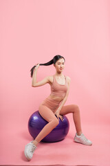 Fit young woman in stylish sportswear posing playfully with an exercise ball against pink backdrop
