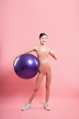 Energetic young woman in athletic wear holds a purple exercise ball smiling against pink background