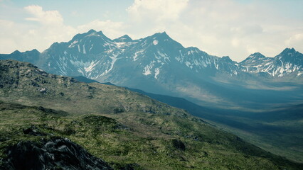 Fototapeta premium Snow capped peaks tower majestically above a vast green valley. Soft clouds drift peacefully across the horizon while sunlight bathes the landscape in warmth and beauty.