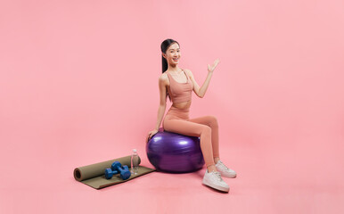 Young woman smiling sitting on balance ball with workout gear on pink background Fitness time