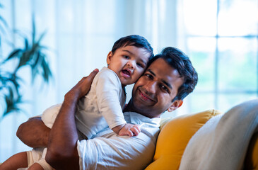 Indian Father and smiling infant boy cuddling on cozy sofa in sunlit modern living space