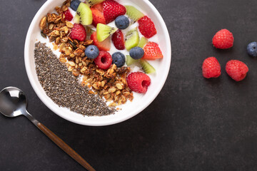 Top view of a white bowl with kefir, granola, chia seeds and fresh fruits on dark background with spoon and raspberries. Horizontal composition with copy space. Concept of healthy breakfast 