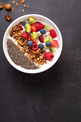 Top view of a bowl with kefir, chia, granola and fresh fruits on dark surface with scattered granola. Copy space. Concept of wholesome breakfast and mindful eating