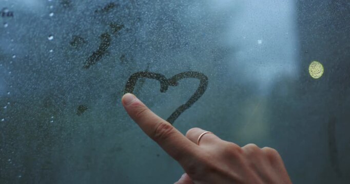 Person draws a heart shape on a foggy, rain-covered car window. Creative, moody, and contemplative moment symbolizing love and connection on a rainy journey.