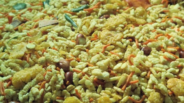 A large, round metal dish is filled with a flavorful Indian snack mix known as namkeen. The golden-colored mixture appears to contain flattened rice, peanuts, and other crispy tidbits.