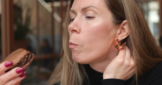 Woman turns profile while biting chocolate macaron sitting in cafe. Lady taster closes eyes and pushes pastry inward touching earring with hand thoughtfully