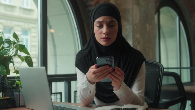 Muslim islamic business woman in hijab typing laptop in office businesswoman receive SMS message with mobile phone reading bad news worried thinking female girl frustrated with smartphone notification