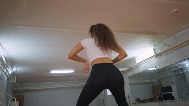 Back view of pretty girl in white top and black leggings twisting body hand on waist during energetic dance session in studio with expressive moves confidence graceful rhythm