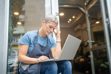 Stressed florist looking at laptop screen with bad business news, worried about flower shop...