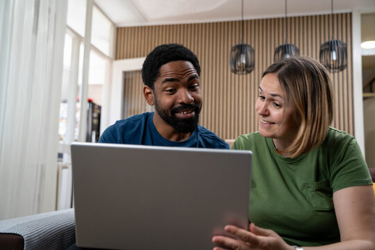 Happy diverse couple smiling and looking at laptop in bright living room during daytime, enjoying love and positive mood. - Powered by Adobe