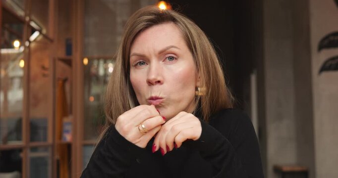 Mature woman sits at cafe table enjoying sandwich during warm conversation. Lady folds hands and taps fingers nodding while listening carefully interlocutor