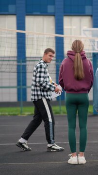 Rear view of young volleyball players standing in sportswear, attentively listening to coach instructing them on an outdoor court, blue building and net in background