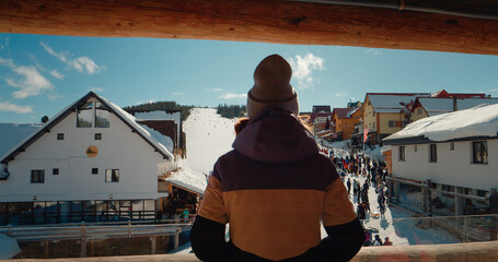 View from the hotel balcony to the snowing ski path resort. Back view of woman dancing while standing on winter resort balcony looking the tourists.