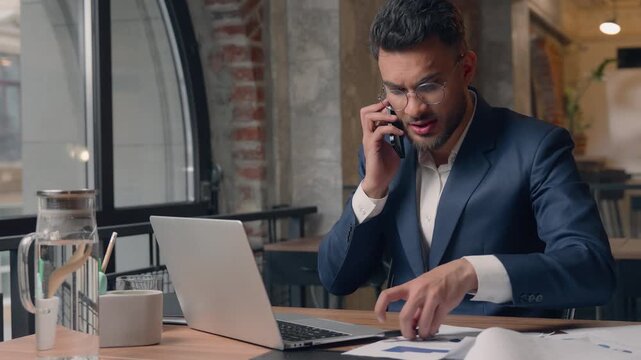 Arabian business male ethnic guy businessman man in office multitask stress busy annoyed Indian agent consultant leader answer mobile phone call talking search data in documents typing laptop online