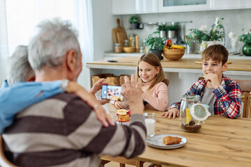 Portret of brother and sister, grandchildren and grandparents having fun together taking a selfie photo with a mobile phone and eating breakfast in kitchen