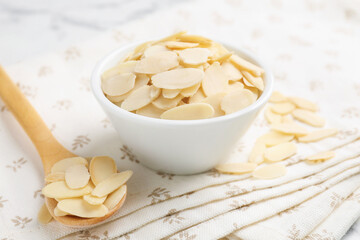 Fresh almond flakes in bowl and spoon on white table, closeup