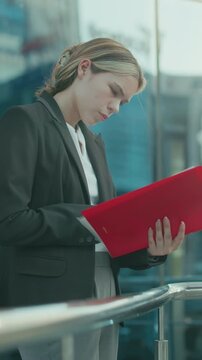 Secretary reading red folder near iron railing outside glass office building reflecting cityscape with cars and sunlight ambiance, showcasing professionalism, focus, and corporate atmosphere