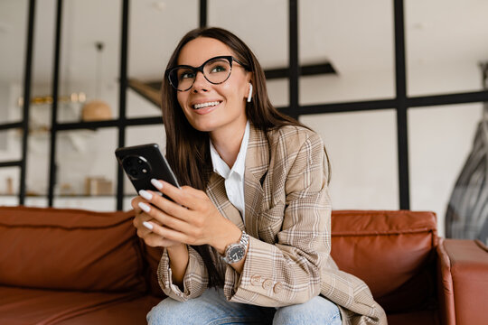 woman sitting on sofa in office relaxed using smartphone in beige jacket and jeans listening to earpods